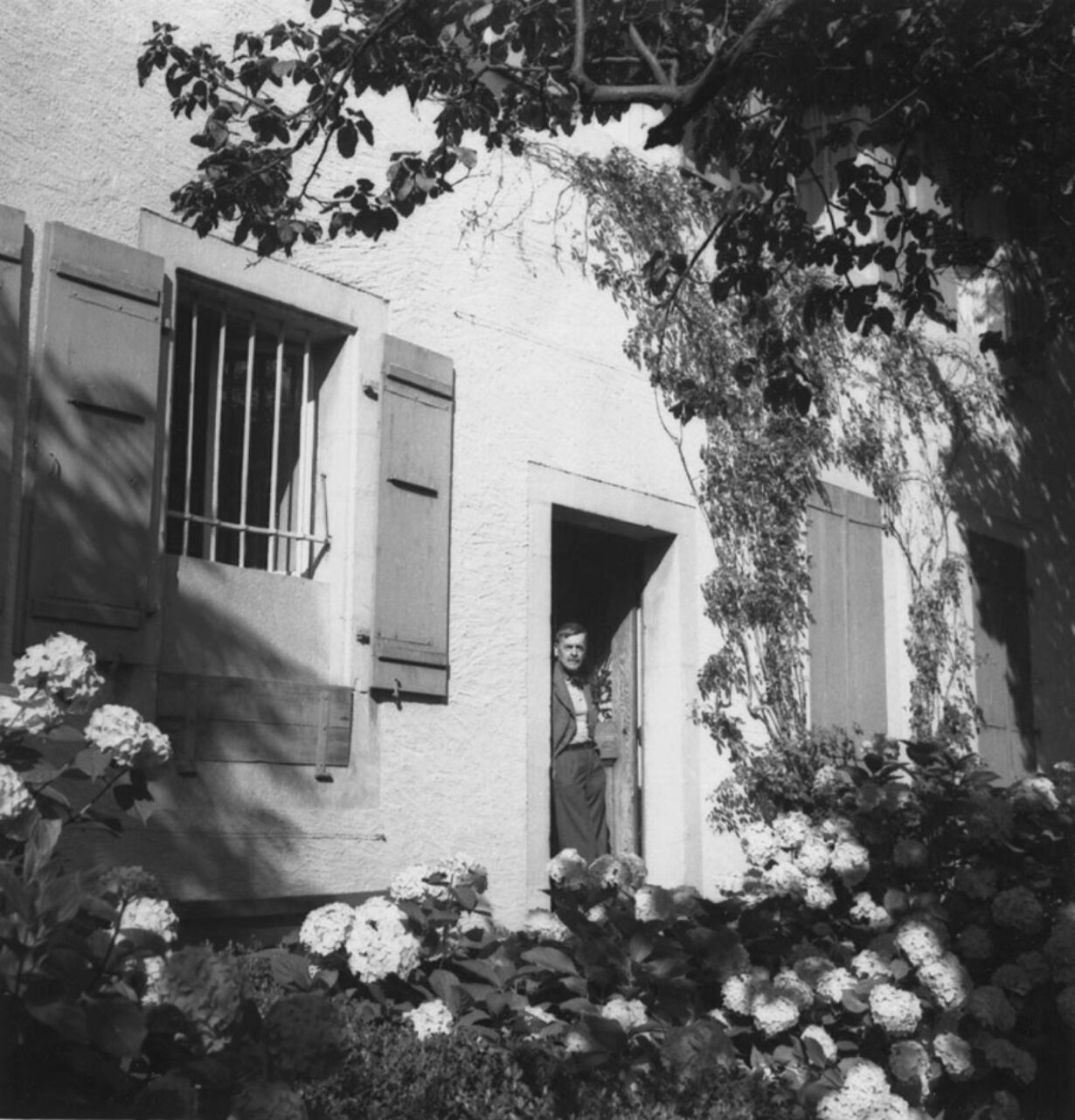 The black-and-white photo shows the garden façade of La Muette, Ramuz's home. The walls are bright, partly covered with ivy and bathed in sunlight. Ramuz is standing in the doorway, his hands in his pockets. The foreground is filled with flowers and tree branches.