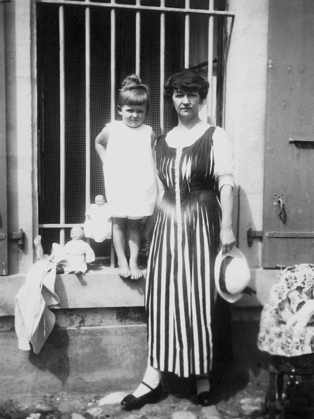 Auf der Fotografie blicken Cécile Cellier-Ramuz und ihre Tochter Marianne in die Kamera. Cécile steht vor einer Hausfassade. Sie trägt ein vertikal gestreiftes Kleid und hat einen weissen Hut in der Hand. Die kleine Marianne steht auf dem Rahmen eines vergitterten Fensters, trägt ein kurzes weisses Kleid und ist barfuss. Neben ihr sitzen zwei Puppen, die ebenfalls weiss gekleidet sind.