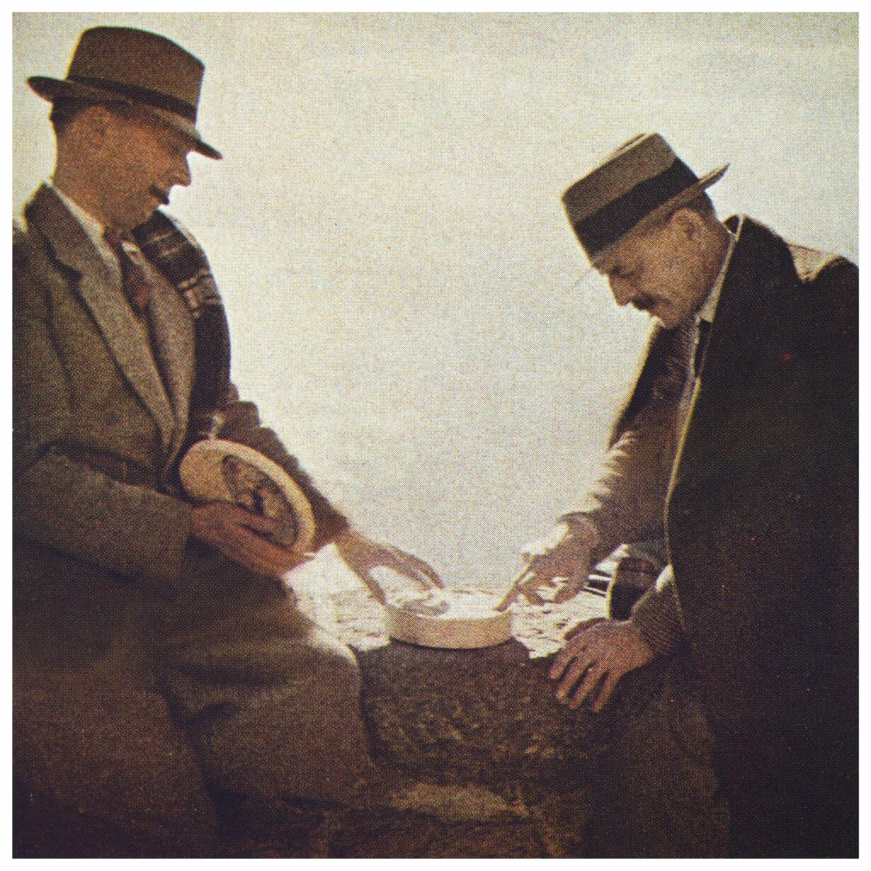 This color photograph shows C.F. Ramuz and Henry-Louis Mermod in Lavaux. They are sitting on a low wall, looking at and touching with their fingertips a vacherin cheese placed between them. Both are wearing hats.