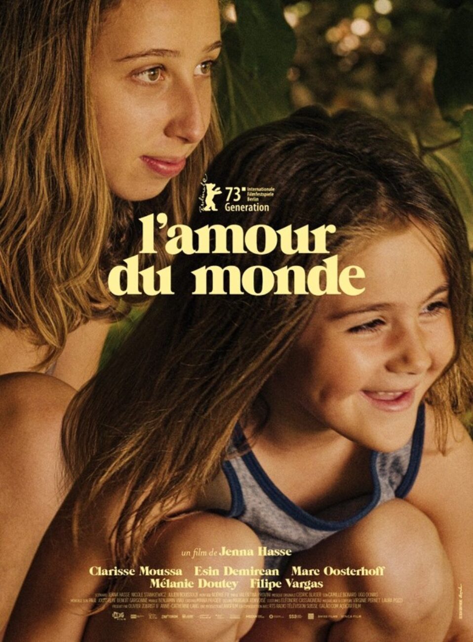 The poster for Jenna Hasse’s L’amour de monde (The Love of the World) features a close-up of a smiling, crouching child. Just behind her is another, slightly older child. Trees can be made out behind them.