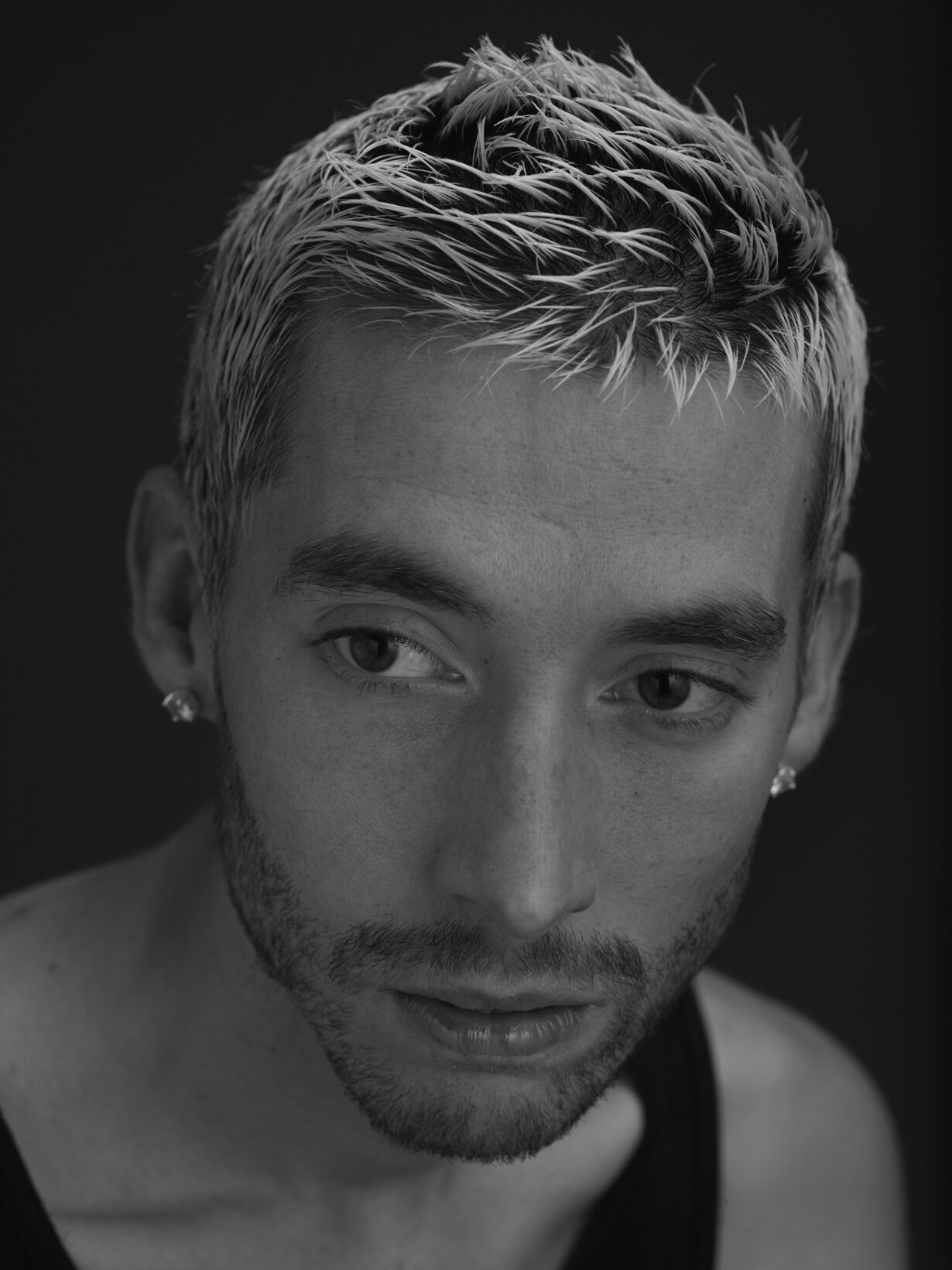 The image is a black-and-white portrait of a young man. He is photographed slightly from above, looking to the left. The tips of his hair are dyed blond.