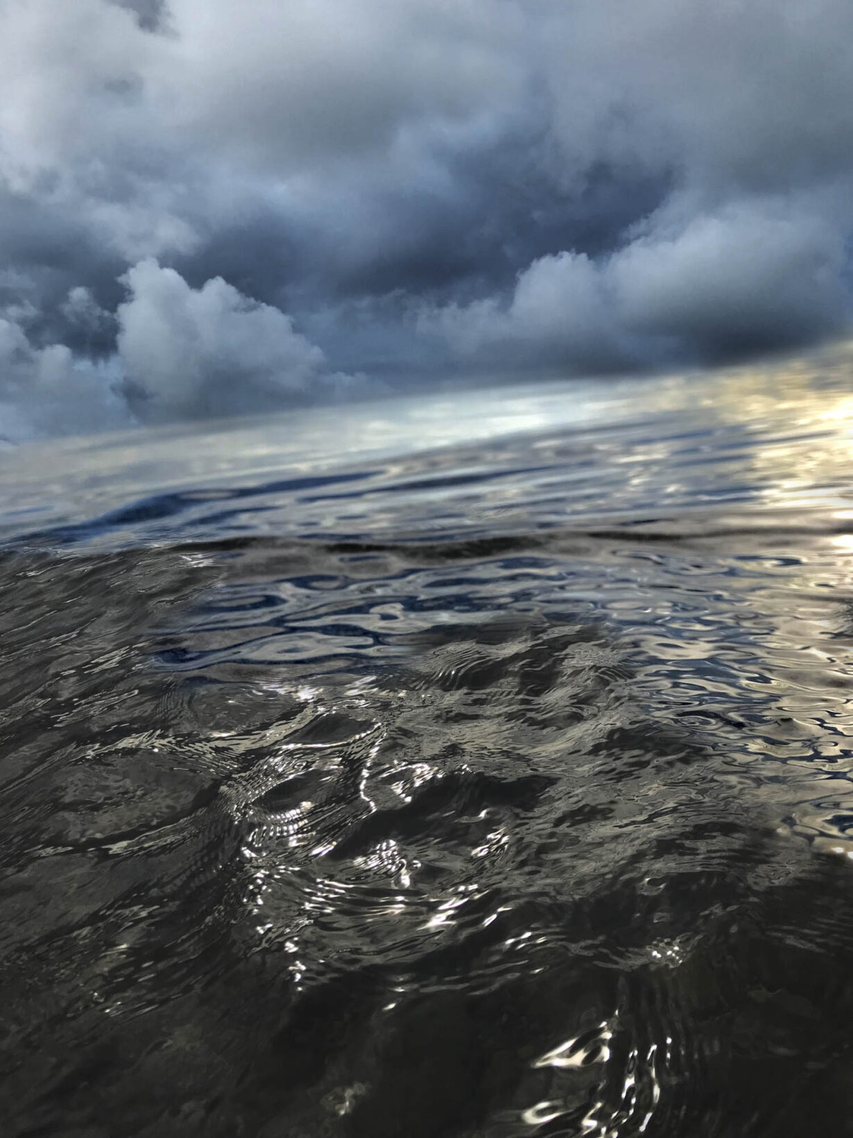 La photo est occupée aux trois quarts par l'eau du lac, vue de très près. L'horizon est penché. Au-dessus de l'eau sur laquelle se reflète la lumière du soleil, nous voyons le ciel saturé de nuages.