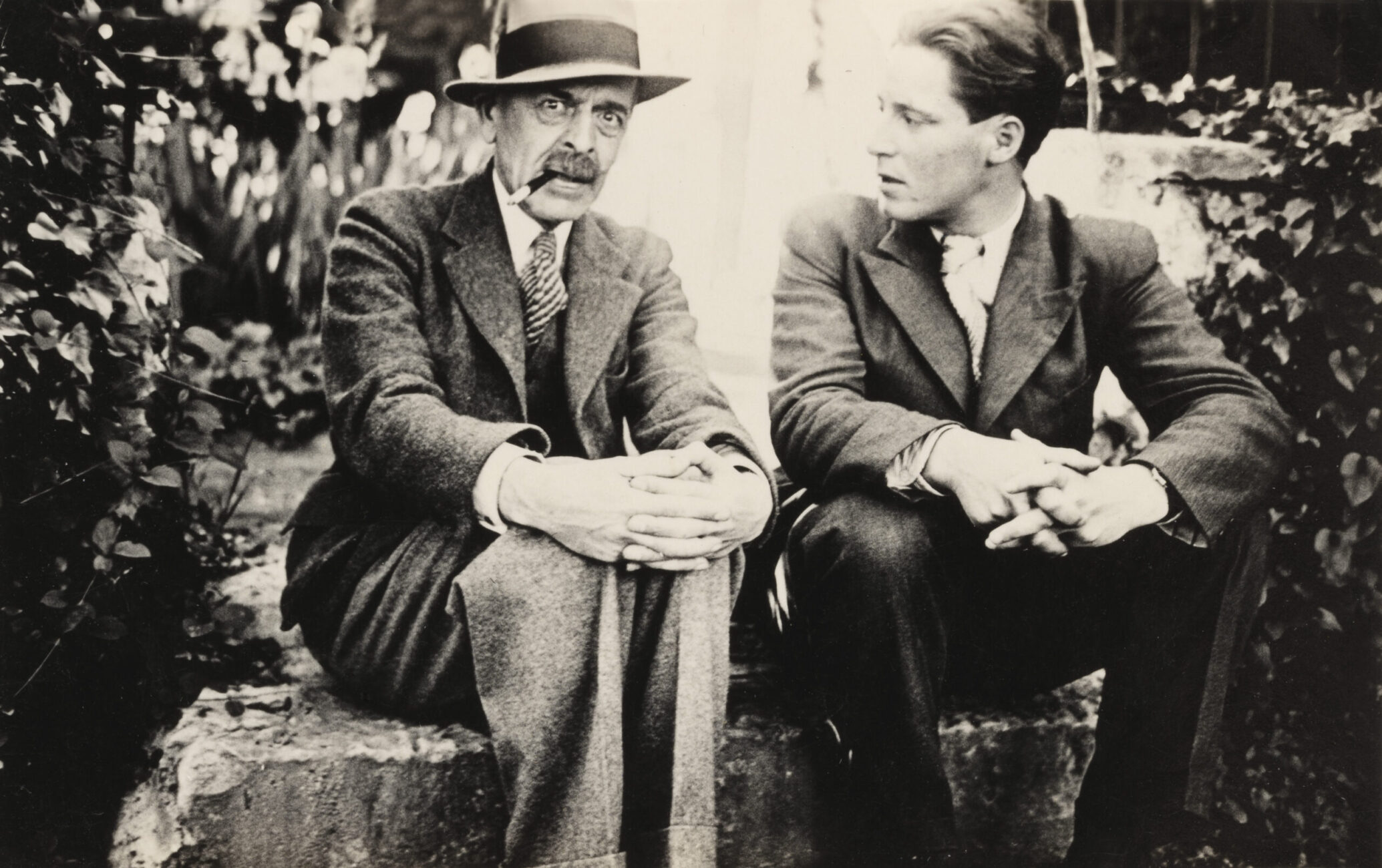 Au centre de la photo en noir et blanc, C.F. Ramuz et Maurice Zermatten sont assis sur une marche en pierre. Tous deux ont les doigts entrelacés sur leurs genoux. Zermatten regarde Ramuz qui regarde l'objectif. Ils sont entourés d'arbres et de plantes.