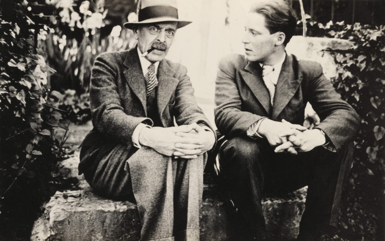 In the center of the black-and-white photo, C.F. Ramuz and Maurice Zermatten are seated on a stone step, their fingers interlaced on their knees. Zermatten is looking at Ramuz, who is looking at the lens. They are surrounded by trees and plants.