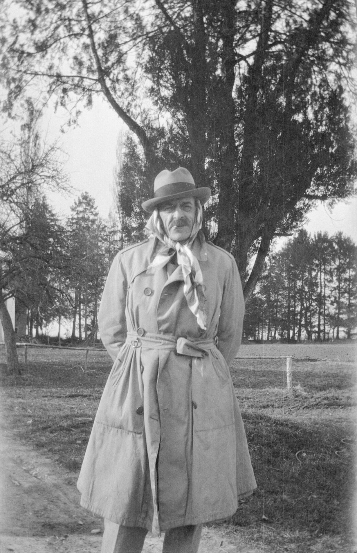 Photo noir blanc de Ramuz debout dans un jardin. Il est emmitouflé dans son imperméable et a entouré sa tête d'un foulard et d'un chapeau.