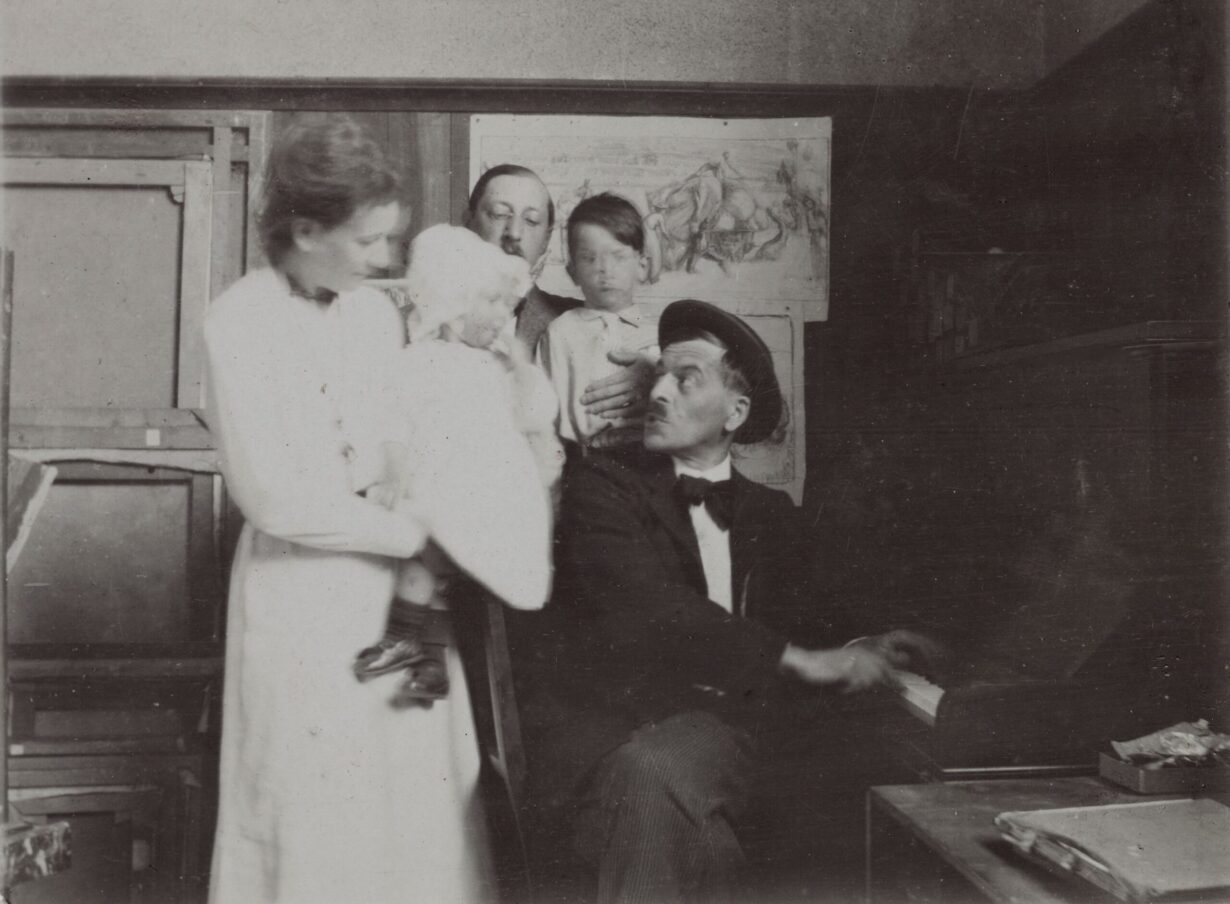 Photo noir blanc de Ramuz jouant du piano pour amuser deux petits enfants dans les bras de deux adultes.