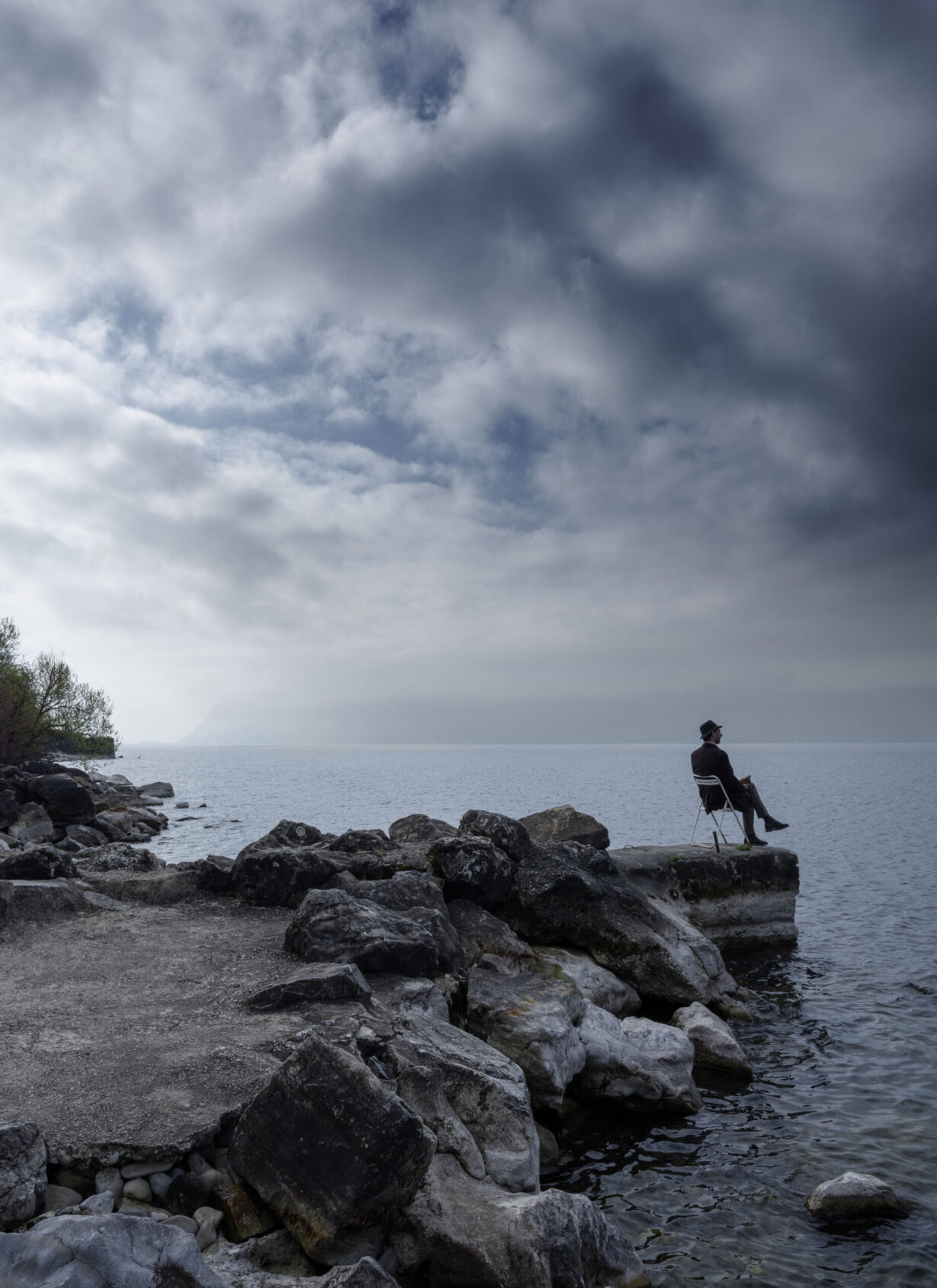Photo contemporaine du bord du lac Léman. Un homme est assis sur une chaise au bout d’une courte jetée.