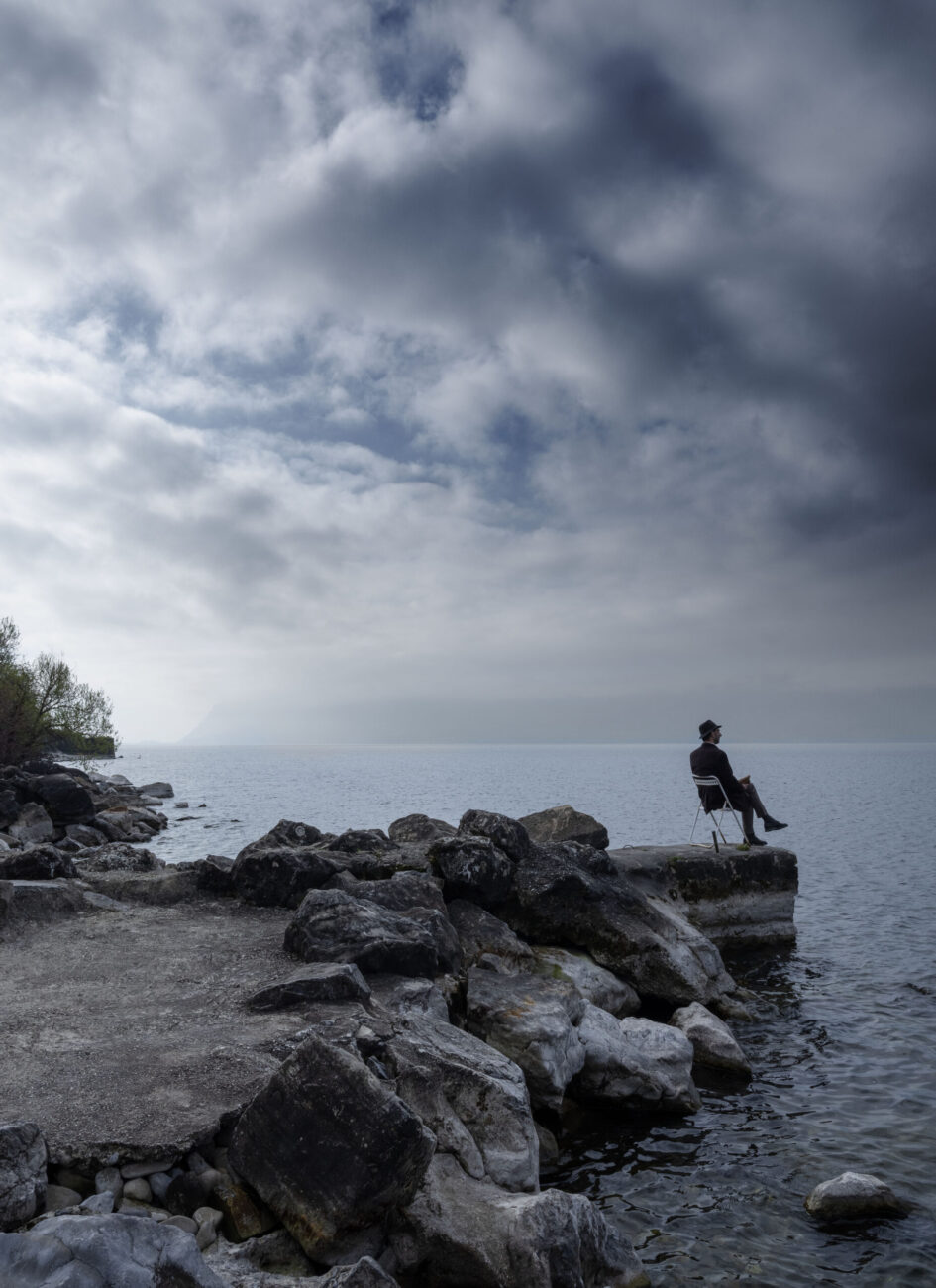 Photo contemporaine du bord du lac Léman. Un homme est assis sur une chaise au bout d’une courte jetée.